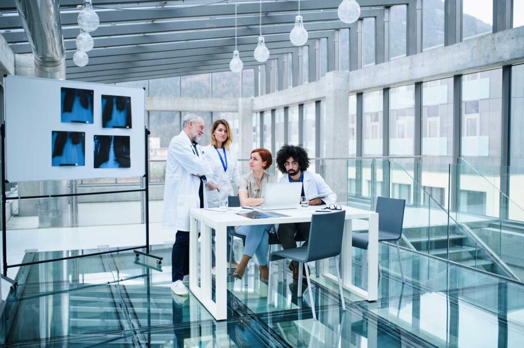 Gruppo di medici in una sala moderna e luminosa, discutono davanti a un tavolo con computer portatile e lastre radiografiche esposte su una lavagna
