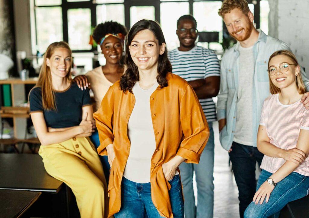 Gruppo di giovani colleghi sorridenti in un ufficio moderno, con una donna al centro che indossa una giacca arancione e posa con sicurezza.