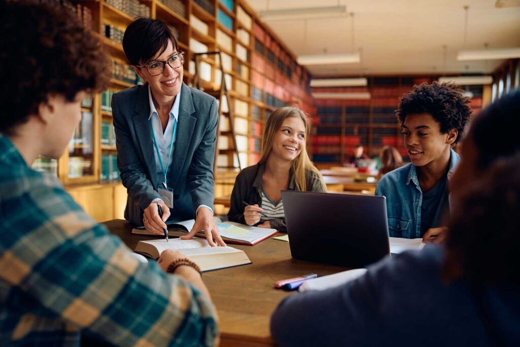 Insegnante sorridente che aiuta un gruppo di studenti delle superiori in biblioteca, con libri aperti e un computer portatile sul tavolo.