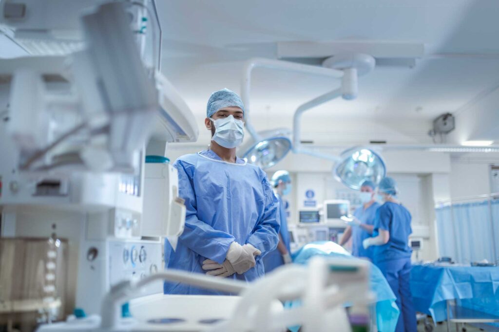 Infermiere specialista in sala operatoria, in piedi con camice sterile e mascherina, mentre altri membri del team medico lavorano sullo sfondo.