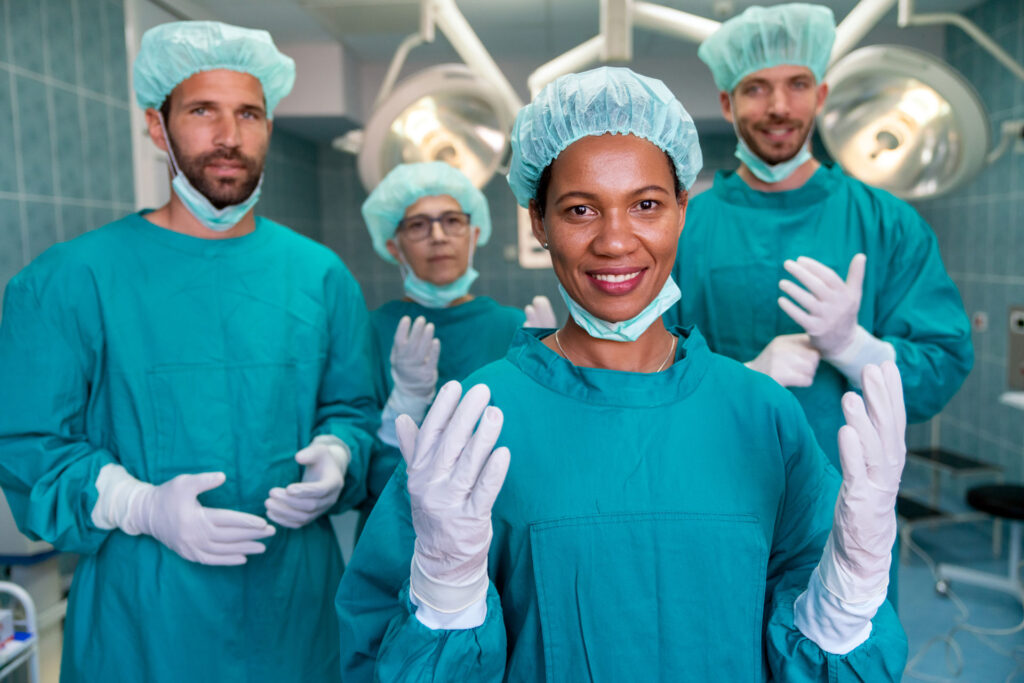 Team di chirurghi in sala operatoria, indossano camici verdi, cuffie e guanti sterili, pronti per un intervento chirurgico, con una donna chirurgo in primo piano che sorride.