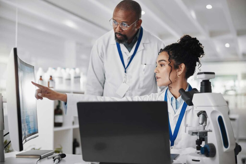 Due scienziati in laboratorio, una donna indica uno schermo del computer mentre un collega osserva; sul tavolo ci sono un microscopio e un laptop.